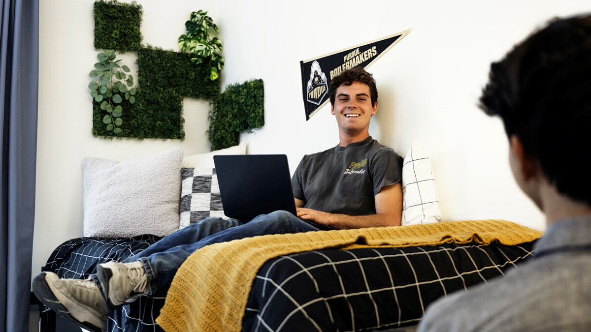 Photo of student sitting on his decorated bed, talking to a friend who's back is to the camera at a residence at Purdue in Indianapolis.. 