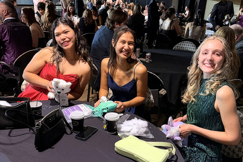 Students in dresses sit around a table and work on creating stuffed animals. 