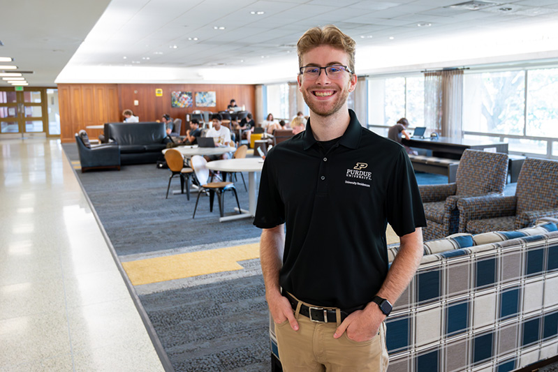 A resident assistant in a Purdue polo smiles while standing in the lobby of a residence hall. 